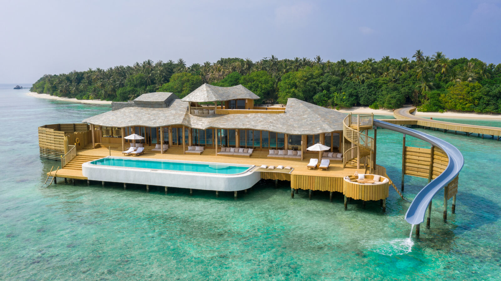 An overwater villa with a pool and slide above clear turquoise water in a tropical setting, the Maldives luxury resort Soneva Fushi Villas offer an idyllic escape. The villa is connected to an island with lush greenery and coconut trees, all under a clear blue sky.