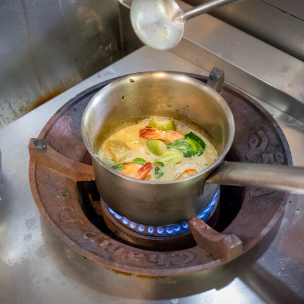 A pot of soup simmering on a stove as part of a Thailand Exclusive Experience at Soneva. The soup features various ingredients, including chunks of vegetables like bell peppers and leafy greens. A metal ladle rests on the edge, ready for stirring or serving in this unique culinary journey.