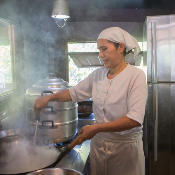 A person in a white chef&#039;s uniform and headscarf is cooking in a kitchen filled with steam at Soneva, Thailand. They are stirring a large pot on a stove, with stacked metal steamers next to them. The kitchen has wooden elements and natural light coming from the windows, adding to the exclusive experience.