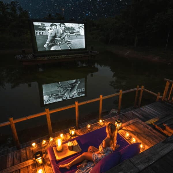 A woman in a summer dress sits on a dock surrounded by lanterns, watching a black-and-white movie on a screen across a small lake. The night sky above is filled with stars, reflecting on the calm water, creating a serene and picturesque outdoor cinema scene—an exclusive Thailand experience by Soneva.