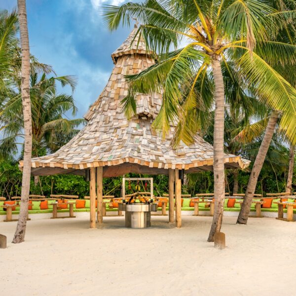 A tropical setting with a wooden gazebo on a sandy beach, shaded by tall palm trees. The gazebo has a unique, layered roof made of wooden shingles, and seating arrangements are visible around it. The sky is partly cloudy, enhancing the serene atmosphere of this Maldives exclusive experience.