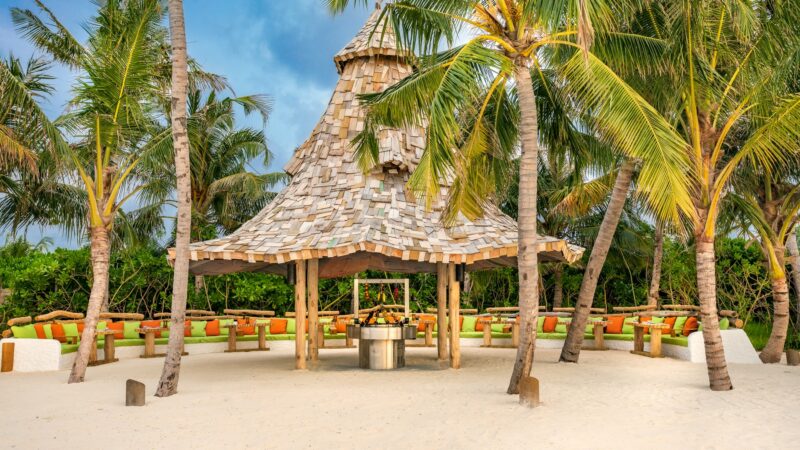 A tropical setting with a wooden gazebo on a sandy beach, shaded by tall palm trees. The gazebo has a unique, layered roof made of wooden shingles, and seating arrangements are visible around it. The sky is partly cloudy, enhancing the serene atmosphere of this Maldives exclusive experience.