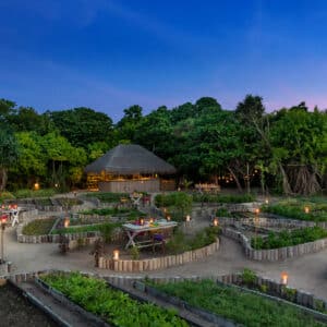 A beautifully lit, lush garden with winding pathways and raised garden beds at dusk evokes the tranquil ambiance of a Maldives luxury resort. Small tables with lit lanterns are scattered throughout, and a thatched-roof building is nestled among the trees in the background under a clear, darkening sky.