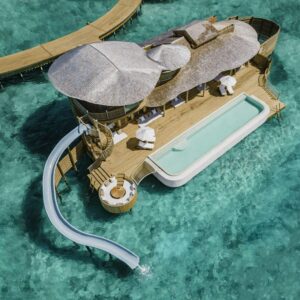 Aerial view of a luxurious over-water villa with a thatched roof, situated in the clear turquoise waters of Soneva Maldives. The villa features a private swimming pool, sun loungers, and a waterslide leading directly into the ocean. A wooden pathway connects it to the land for an unforgettable holiday.