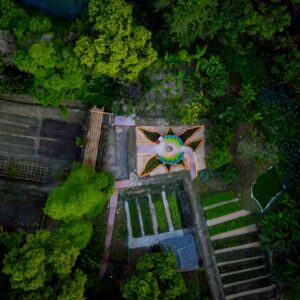 Aerial view of a lush green forest surrounding a clearing at Soneva Kiri Resort, where geometric garden plots and a central, star-shaped structure captivate the eye. The garden plots are arranged in neat rows, with pathways gracefully leading to the flower-shaped design at the center.