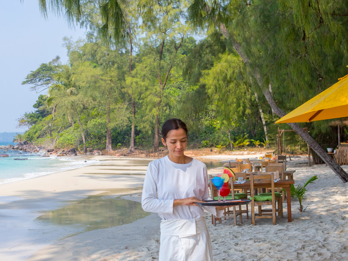 A server in a white uniform holds a tray with colorful drinks on a pristine, sandy beach at Soneva in the Maldives. Surrounded by lush greenery and a calm sea, wooden tables and chairs are set up under a canopy along the shoreline, creating a serene outdoor dining area perfect for celebrating Chinese New Year.