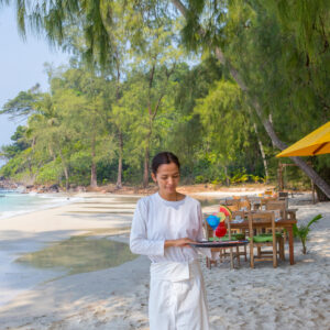 A server in a white uniform holds a tray with colorful drinks on a pristine, sandy beach at Soneva in the Maldives. Surrounded by lush greenery and a calm sea, wooden tables and chairs are set up under a canopy along the shoreline, creating a serene outdoor dining area perfect for celebrating Chinese New Year.