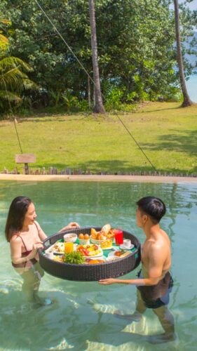 A woman and a man enjoy a floating breakfast in a pool surrounded by tropical greenery. The woman is in a bikini, the man in swim trunks, as they stand in the water holding a large tray filled with assorted food and drinks. This Thailand exclusive experience offers stunning views of the beach and sea beyond.