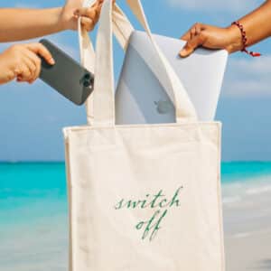 Two hands place a phone and laptop into a cream-colored tote bag with "switch off" written on it, perfect for those seeking Soneva Exclusive Offers. In the background, the serene beach displays turquoise water and a clear blue sky.