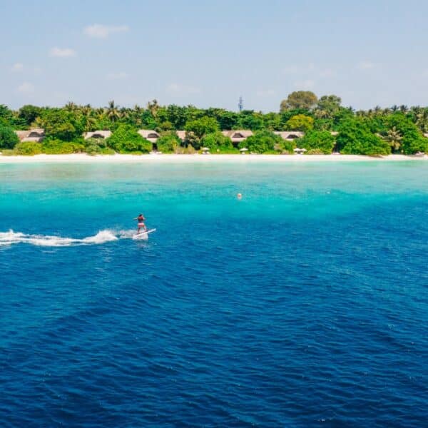 A person rides a jet ski on the bright blue waters near a tropical island with lush green trees and several beach huts. The sky is clear with a few clouds, and the beach in the background features white sand—a true Maldives exclusive experience at Soneva.