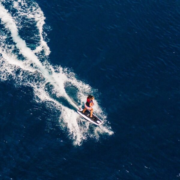 A person is riding a jet ski on a large body of water, leaving a white wake trail behind them. The deep blue water reflects the opulence of a Maldives luxury yacht holiday, and the scene is captured from an aerial perspective.