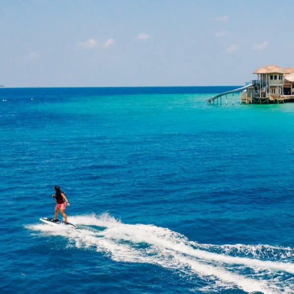 A person rides a jet ski on the vibrant blue ocean, leaving a white trail behind. In the background, there is a luxurious overwater villa with straw roofs on stilts, part of the Maldives&#039; exclusive experience at Soneva. The sky is clear with a few scattered clouds.