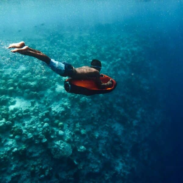 A person in swim trunks is underwater, using an underwater scooter to navigate through a coral reef. Sunlight filters through the clear blue water, illuminating the coral and creating a serene aquatic scene—truly a Maldives exclusive experience at Soneva.