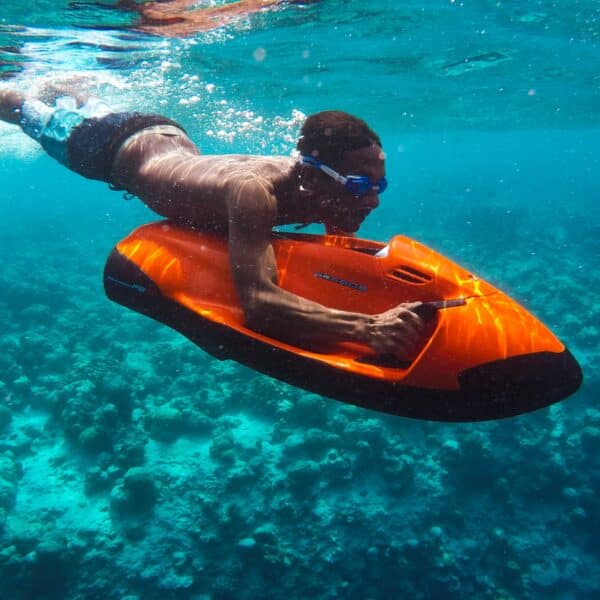 A person wearing swim goggles is underwater, gripping an orange and black motorized scooter, propelling themselves through clear blue water above a coral reef. This Maldives exclusive experience at Soneva showcases vibrant marine life and colorful coral structures in a breathtaking underwater scene.