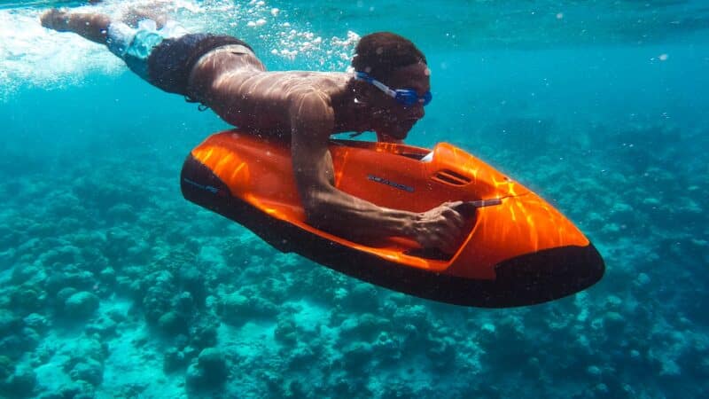 A person wearing swim goggles is underwater, gripping an orange and black motorized scooter, propelling themselves through clear blue water above a coral reef. This Maldives exclusive experience at Soneva showcases vibrant marine life and colorful coral structures in a breathtaking underwater scene.