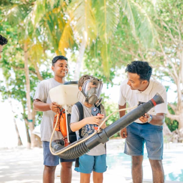 A group of three men watches as a young boy, wearing a large mask, tries on a backpack sprayer. They are outdoors at an exclusive Soneva resort in the Maldives, surrounded by lush green trees, with a pool in the background. The scene is lit with natural sunlight.