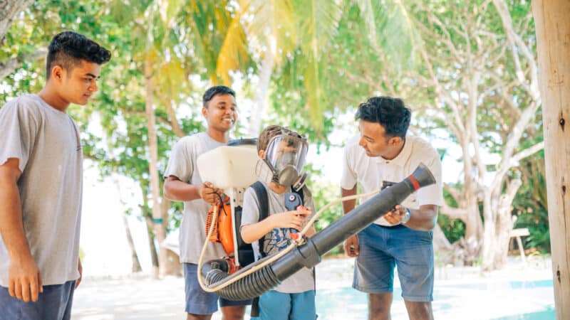 A group of three men watches as a young boy, wearing a large mask, tries on a backpack sprayer. They are outdoors at an exclusive Soneva resort in the Maldives, surrounded by lush green trees, with a pool in the background. The scene is lit with natural sunlight.