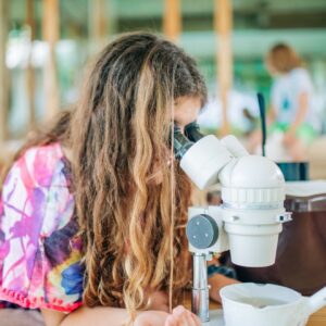 A young girl with long brown hair uses a microscope on a wooden table. She wears a colorful tie-dye shirt. Other children, blurred in the background, are sitting and engaged in different activities in a bright and spacious room, part of an initiative supported by the Soneva Foundation.