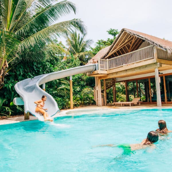 A child slides down a water slide into a tropical pool, part of the exclusive Soneva experience in the Maldives, while two other children swim nearby. The pool is surrounded by lush greenery and palm trees, with a thatched-roof house visible in the background.