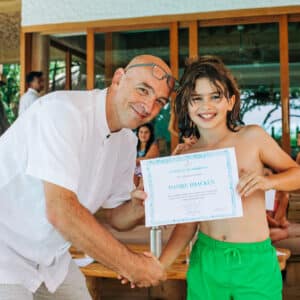 A smiling man in a white shirt shakes hands with a young boy in green shorts. The boy, holding up a certificate with both hands, seems thrilled. They are indoors with family sitting in the background, possibly reminiscing about their recent holiday at Maldive Soneva. The atmosphere appears celebratory.