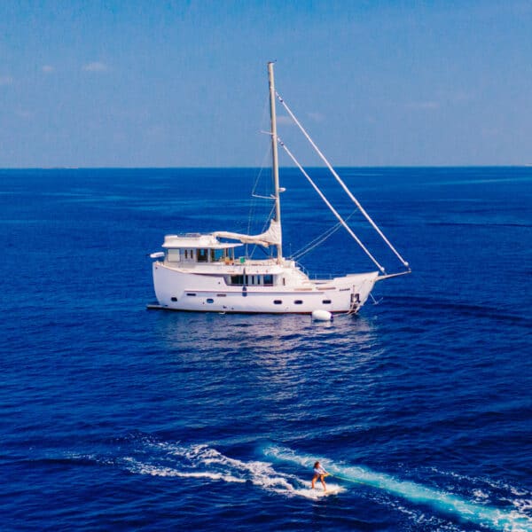 A white sailboat floats on a vast, calm blue ocean under a clear sky. In the foreground, a person rides a jet ski, leaving a trail of white water on the otherwise serene sea. The horizon is distant and faint, capturing the essence of a Maldives Luxury Yacht Holiday at Soneva.