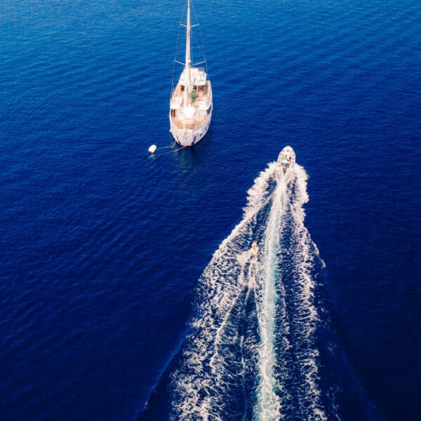 Aerial view of a sailing boat anchored in the deep blue sea during a Maldives luxury yacht holiday at Soneva, with a smaller motorboat speeding away, leaving a trail of white waves behind. The calm, expansive ocean contrasts with the dynamic motion of the motorboat.