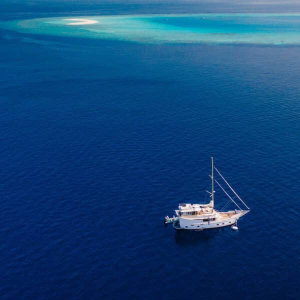 A luxury yacht floats on calm, deep blue ocean waters with a distant sandbar visible in the background. The clear, turquoise waters near the sandbar contrast with the deep blue of the surrounding sea, creating a serene and picturesque scene perfect for a Maldives holiday.