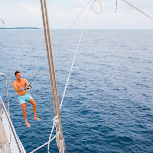 A man in blue swim trunks swings out over the ocean from the side of a luxurious sailboat on a rope. The sky is slightly cloudy, and the expansive blue ocean stretches out in the background. Enjoying his Maldives Luxury Yacht Holiday at Soneva, the deck and some rigging are visible.