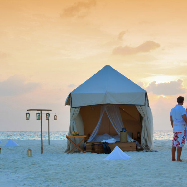 A couple stands arm in arm on a sandy beach at sunset, facing a large, open white tent. The tent is furnished with a bed and surrounded by lanterns on the sand. Small white pyramid-shaped objects and a stand with more lanterns decorate the beach, offering an exclusive experience reminiscent of Soneva's Maldives retreats.