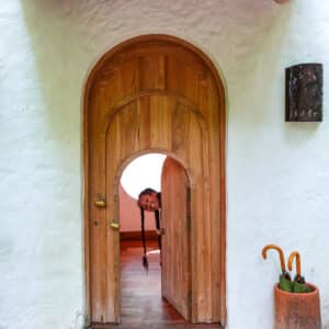 A wooden doorway within a white stucco structure features a smaller door beneath the main one. Both are open, revealing a person peeking through the smaller door. A pair of umbrellas are placed in a cylindrical holder to the right, reminiscent of an inviting entrance at a family holiday in Soneva, Maldives.