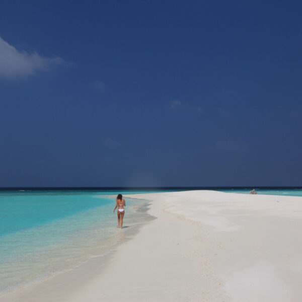 A solitary person walks along a pristine, white sandy beach that stretches into the distance, bordered by vibrant turquoise water on a clear, sunny day with a deep blue sky. The tranquil scene exudes serenity and natural beauty, embodying the Maldives Exclusive Experience at Soneva resorts.