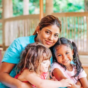 A smiling woman in a blue shirt sits on the floor and hugs two young girls, one with dark hair in pigtails wearing a pink shirt, and the other with light brown hair wearing a red dress. They are in a bright room with wooden floors, reminiscent of their recent family holiday at Soneva in the Maldives.