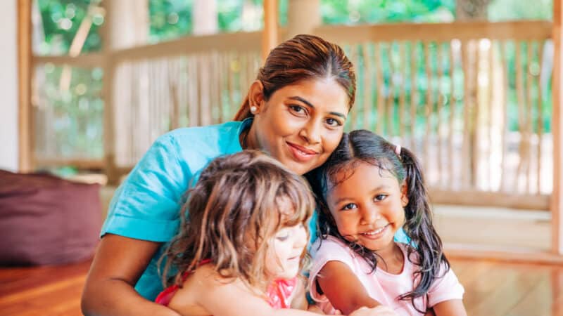 A smiling woman in a blue shirt sits on the floor and hugs two young girls, one with dark hair in pigtails wearing a pink shirt, and the other with light brown hair wearing a red dress. They are in a bright room with wooden floors, reminiscent of their recent family holiday at Soneva in the Maldives.