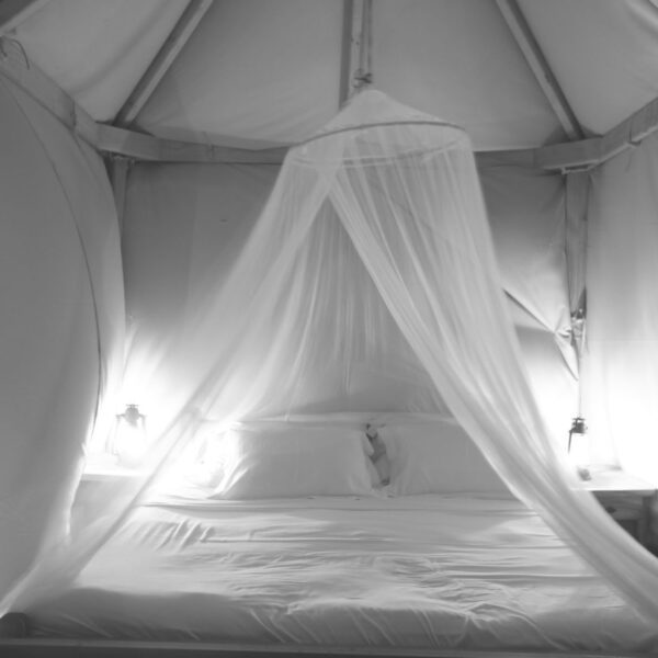 A cozy bed inside a white tent with a mosquito net draped over it. Two lanterns are placed on either side of the bed, casting a soft glow. The setting exudes a calm and serene atmosphere, perfect for an exclusive Soneva experience or a peaceful retreat in the Maldives.