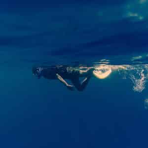 A person wearing snorkeling gear swims near the surface of the clear blue water during a Maldives exclusive experience. Light filters through the water above, creating a serene underwater scene. Air bubbles trail behind the swimmer, adding a dynamic touch to this tranquil Soneva paradise.