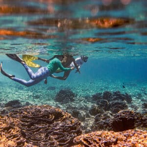 A person wearing snorkeling gear swims underwater near a vibrant coral reef during a family holiday at Maldives&#039; Soneva resort. The clear water reveals the colorful coral formations below, while sunlight from above creates a serene and visually striking scene.