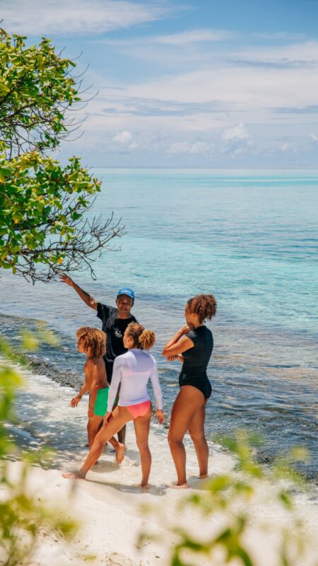 A family of four stands on a sandy beach by clear turquoise waters, enjoying their Maldives exclusive experience. An adult points towards the ocean, while the other adult and two children listen attentively. The sky is clear, and a leafy tree branch extends into the scene from the left.