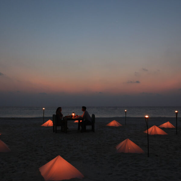 A couple sits at a table enjoying a romantic dinner on the beach at dusk. The scene is illuminated by warm candlelit lanterns arranged in the sand around them, creating a cozy and intimate atmosphere. The sky is a gradient of darkening twilight colors, epitomizing the Maldives' exclusive experience.