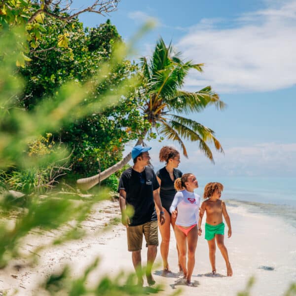 A family of four, dressed in casual beachwear, stands on a sandy shore lined with lush greenery and palm trees at Soneva in the Maldives, gazing at the ocean under a clear blue sky. The scene suggests an exclusive, peaceful, sunny day at the beach.