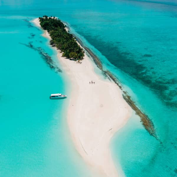 Aerial view of a narrow, white sandy island surrounded by turquoise water. The island, reminiscent of a Maldives exclusive experience, is covered with green vegetation on one end. A small boat is anchored near the shoreline while a few people enjoy walking along the beach.