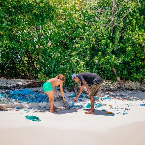 Two people clean up debris on a sandy beach near dense greenery. They are gathering scattered pieces of blue and green fishing nets. The beach, partially shaded by trees, suggests an environmental conservation effort as part of a Maldives Exclusive Experience at Soneva.