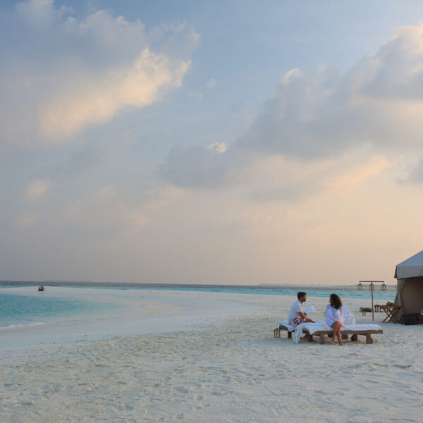 A couple sits on lounge chairs by a table under a tent on a pristine beach with white sand and turquoise water, beneath a partly cloudy sky. The tranquil scene suggests an exclusive Soneva getaway in the Maldives, offering a serene and secluded experience.