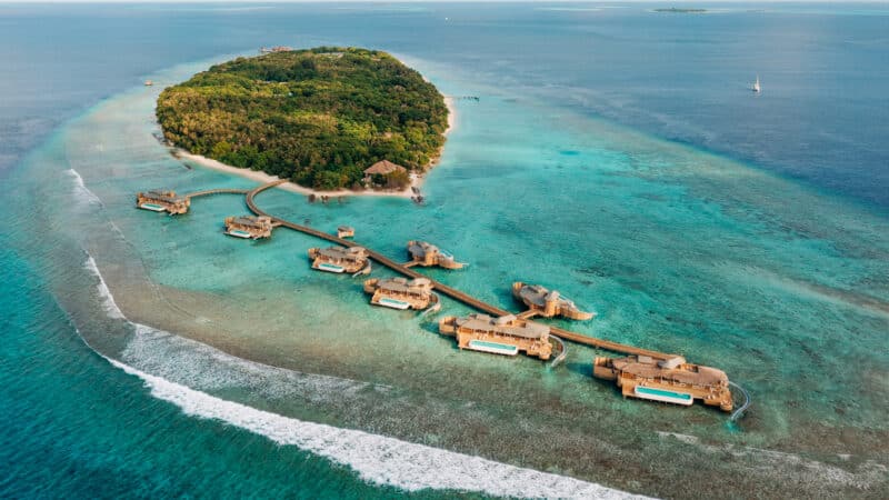 Aerial view of an island resort surrounded by blue waters. The lush island, connected by wooden walkways to overwater bungalows extending into the sea, looks like paradise. A sailboat is visible in the distance as waves break near the shore. Discover Soneva Exclusive Offers for a luxurious getaway.