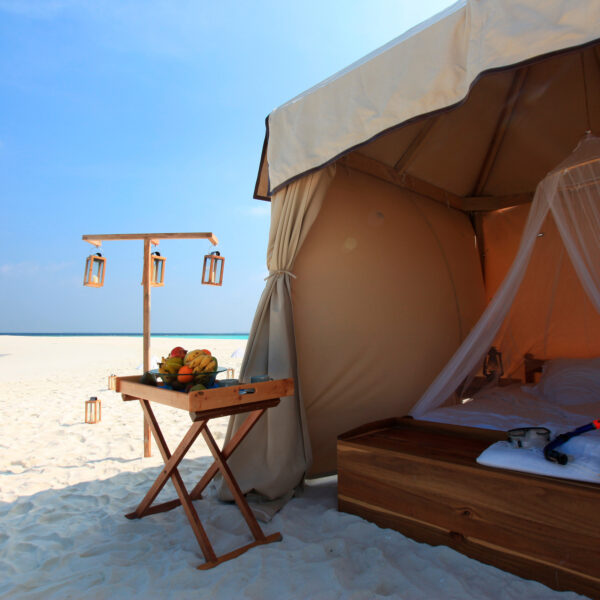 A luxurious tent set up on a pristine sandy beach with a clear blue sky and turquoise ocean in the background. Inside the tent is a cozy bed with mosquito netting. A nearby wooden table holds fresh fruit and drinks. Lanterns hang on a wooden frame next to the tent, offering the ultimate Maldives exclusive experience.