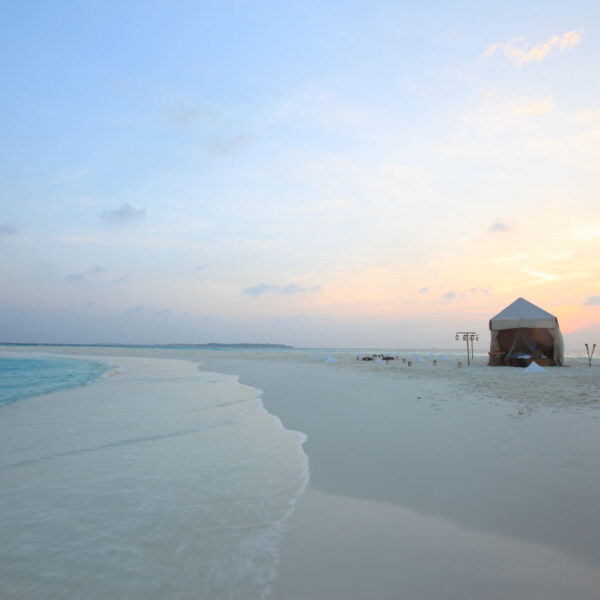 A serene beach scene at sunset, with soft waves gently lapping against the white sandy shore. A small, simple tent sits alone on the sand, with a couple of lounge chairs nearby. The sky is painted in pastel hues of blue, pink, and orange—a true Maldives Exclusive Experience by Soneva.