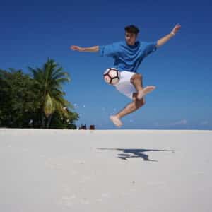 A person wearing a blue shirt and white shorts jumps and performs a skillful kick with a soccer ball on a sandy beach during their family holiday at Soneva in the Maldives. Palm trees, beach chairs, and clear blue sky are in the background, while the person's shadow is visible on the sand.