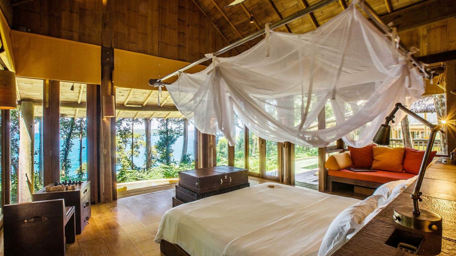 A tropical bungalow bedroom with a large canopy bed draped in white mosquito netting, perfect for a Thailand family holiday. Wooden walls and floors, large windows offering views of lush greenery and the ocean, and a cozy seating area with red and orange cushions complete this Soneva-style paradise.