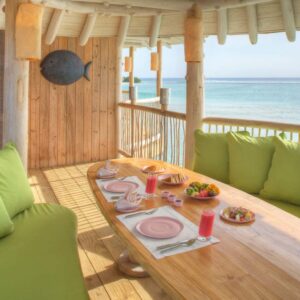 A tropical dining area at a Maldives luxury resort features a wooden table set for a meal on a balcony overlooking the ocean. The table, adorned with Soneva's finest place settings, includes a pink drink and various plates of snacks. Lime green cushions provide seating, and a bedroom is visible through the doorway.