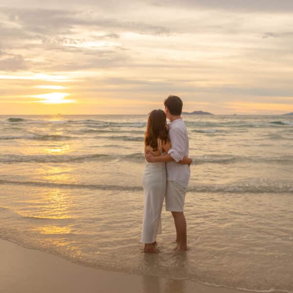 A couple stands on the shore, embracing and watching the sunset over the ocean. Dressed in white, they revel in the serene beauty of gentle waves, a sandy beach, and a colorful sky with clouds reflecting the setting sun. It&#039;s moments like these that Soneva Exclusive Offers make unforgettable.