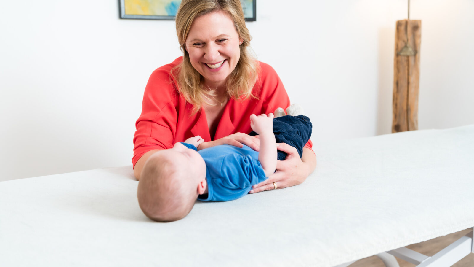 A smiling woman in a red shirt cradles a baby lying on a white table in a bright room. A colorful painting hangs on the wall in the background, and a wooden lamp is nearby.